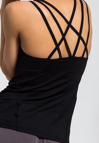 Vue arrière d'une personne portant un débardeur noir avec plusieurs bretelles fines entrecroisées sur les épaules et un bas gris.