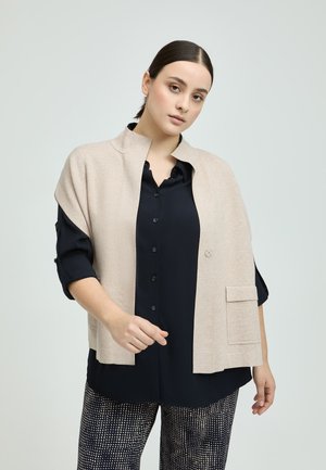 Beige gestrickte Weste mit hohem Kragen, über einem dunkelblauen Hemd mit Knopfleiste, das eine Fronttasche und aufgerollte Ärmel hat.