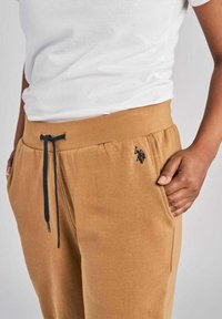 Pantaloni della tuta in cotone marrone con cintura elasticizzata, cordoncino, tasche laterali e un piccolo logo nero sul lato sinistro della coscia.