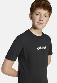 Melns kokvilnas T-krekls ar īsām piedurknēm. Priekšpusē ir balts Adidas logo. Standarta apļa kakla dizains, gluda tekstūra.