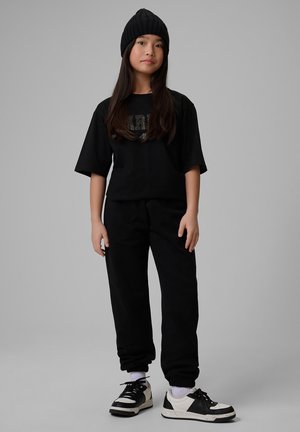 Dziewczyna z długimi ciemnymi włosami nosi czarną czapkę, czarny oversize t-shirt, czarne spodnie, białe skarpetki oraz czarno-białe sneakersy, stoi na szarym tle.