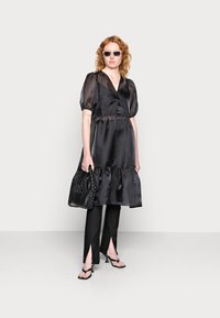 Vestido de cetim preto com mangas bufantes e saia em camadas, combinado com calças skinny pretas e sapatos de salto com tiras. Os acessórios incluem uma bolsa trançada e óculos de sol.