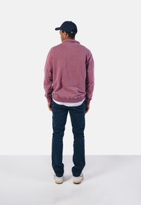 Pull en polaire rose avec un col montant, associé à un pantalon bleu foncé. Des baskets blanches complètent la tenue. La vue est de dos.