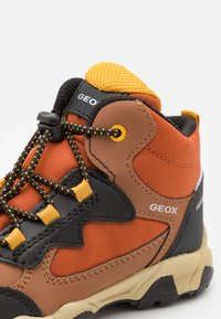 Chaussure de randonnée marron et orange en matériaux mesh et cuir, semelle en caoutchouc, accents jaunes et détails noirs contrastants. Finition texturée.