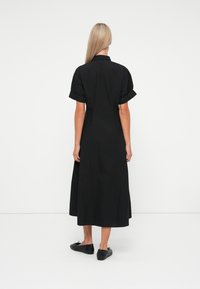 Donna con lunghi capelli biondi che indossa un abito midi nero a maniche corte e scarpe basse nere, in piedi contro uno sfondo bianco, vista da dietro.