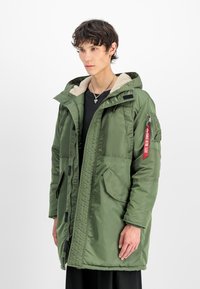 Giacca puffer verde oliva, con cappuccio, foderata in pile, tasche frontali e un'etichetta rossa sulla manica. Finitura liscia e lucida.