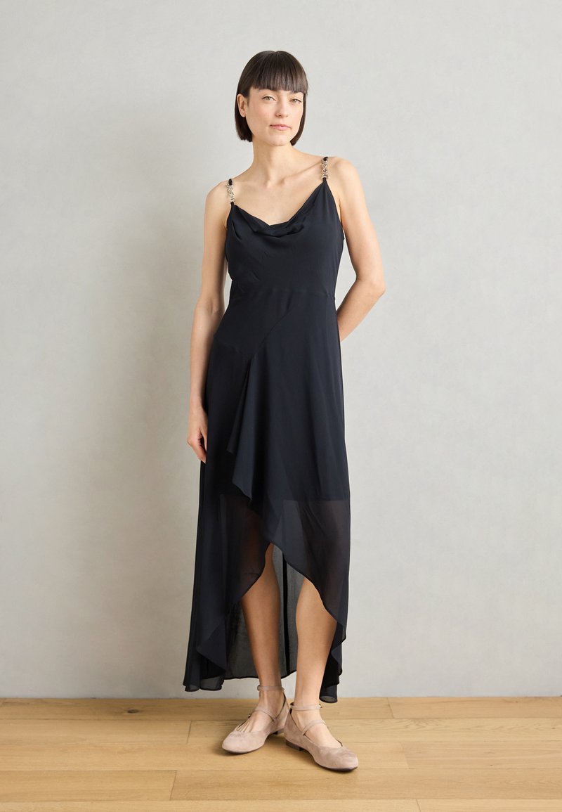 Schwarzes Kleid mit einem Cowl-Ausschnitt und asymmetrischem Saum. Besitzt ein durchscheinendes Material und veredelte Träger. Trägt man zu beigen Ballerinas.