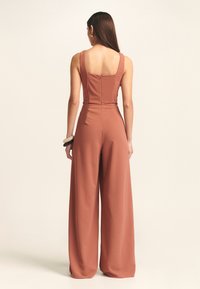 Vrouw met lang haar in een mouwloze roestkleurige jumpsuit met wijdere pijpen, gezien vanachter, staand tegen een effen achtergrond.