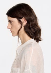 Blouse blanche ample et transparente avec un subtil motif rayé. Modèle avec des cheveux bruns mi-longs, portant de petites boucles d'oreilles en forme de cercle. Vue de profil.