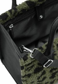 Vista interior de una bolsa de lona con patrón negro y verde que muestra un bolsillo con cremallera, un cierre de metal y una correa de hombro ajustable.
