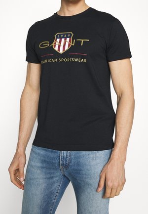Mann trägt schwarzes GANT T-Shirt mit goldenem und rotem Logo und Schriftzug, kombiniert mit blauen Jeans, steht vor weißem Hintergrund.