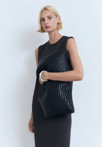 Schwarze, geflochtene Handtasche, gehalten gegen ein dunkles Kleid. Glatte Oberfläche, rechteckige Form und große Größe, geeignet für den Transport mehrerer Gegenstände.