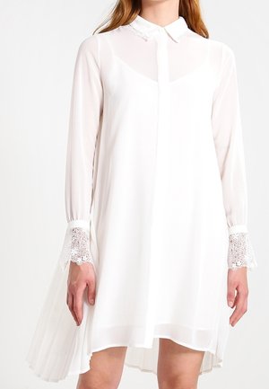 Robe blanche à manches longues en tissu léger. Présente un col, un devant à boutons et des accents en dentelle sur les manches. Détail de ourlet plissé.
