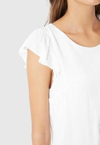 Camiseta blanca de algodón con mangas cortas con volantes y escote redondo. Textura suave con un corte relajado, destacando los detalles de diseño femenino.