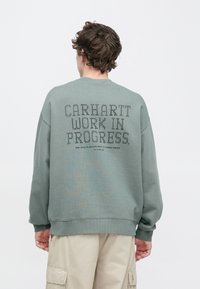 Sudadera verde con cuello redondo, con la inscripción "CARHARTT WORK IN PROGRESS" en negro en la parte trasera. Textura suave, diseño de ajuste relajado.