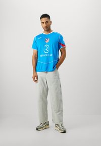 Blue soccer jersey with red accent, white trim, and logo. Paired with light gray cargo pants and patterned sneakers. Simple background.
