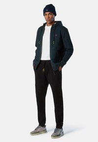 Felpa con zip verde scuro con accenti gialli, t-shirt bianca sotto, pantaloni jogger neri e sneaker grigio-verdi. Abbinato con un berretto blu navy.