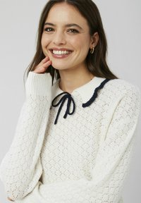 Pull en tricot blanc avec motif en losanges, poignets côtelés et col en forme de vagues bleu marine. Présente un nœud assorti au col.