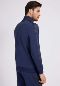 Marineblaues Sportjacke mit hohem Kragen, langen Ärmeln und schwarzen gemusterten Seitenstreifen. Glatte Textur mit gerippten Bündchen und Saum.