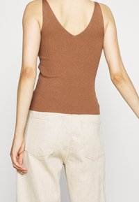 Personne portant un débardeur côtelé marron et un pantalon beige clair, vue de dos avec les bras détendus le long du corps.