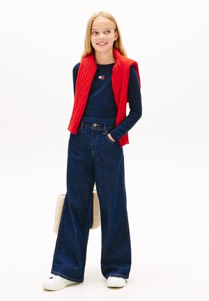 Czerwona pikowana kamizelka na granatowej, prążkowanej koszulce z długim rękawem, do tego szerokie jeansy w ciemnym kolorze i białe trampki. Trzyma kremową torebkę.