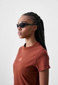 Sunglasses with black cat-eye frames and dark lenses, paired with a short-sleeve, rust-colored t-shirt featuring an orange logo.