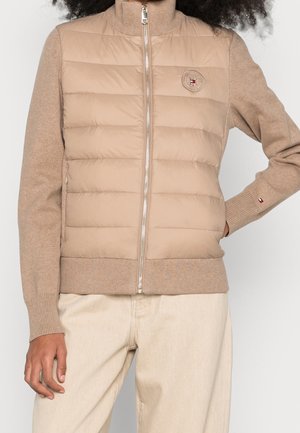 Frau trägt eine beige gesteppte Jacke mit Reißverschluss, Strickärmeln und einem Logo-Patch auf der linken Brust, kombiniert mit hellcremefarbenen Hosen.