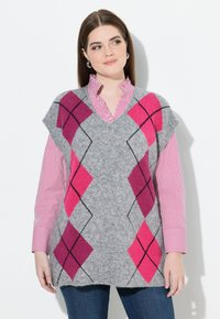Žena v šedé bezrukávové argyle svetr vestě s růžovými a fialovými diamanty přes pruhovanou červenou a bílou blůzu a modré džíny.