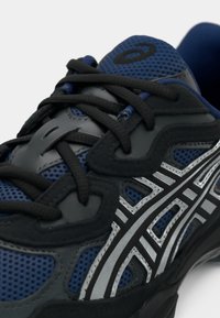 Gros plan d'une chaussure de sport bleue et noire avec des lacets noirs et des détails argentés réfléchissants.