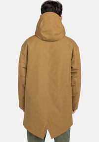 Mann mit dem Rücken zur Kamera, der einen langen, camelbraunen, mit Kapuze versehenen Mantel trägt, der eine zentrale Rückennaht und einen kleinen Schlitz am Saum hat.