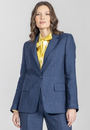 Kobieta z falującymi brązowymi włosami, ubrana w niebieski garnitur i żółtą bluzkę z kokardą, stojąca na tle jasnoszarego tła.