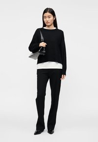Pull noir surdimensionné avec ourlet côtelé, superposé à une chemise blanche. Associé à un pantalon noir sur mesure et un sac à main noir brillant.