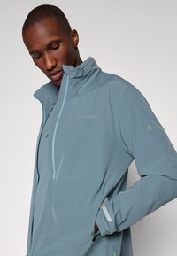 Veste imperméable bleu clair avec un col haut, fermeture éclair et un logo de marque discret sur la poitrine. Présente des accents réfléchissants et des poches zippées.