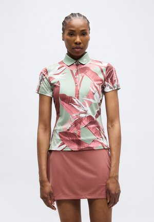 Ženska v polo majici s kratkimi rokavi z zelenim in roza tropskim listnim potiskom ter v enobarvni roza krilu, stoji pred enostavnim ozadjem.