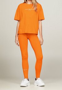 Camiseta naranja con logo, corte holgado; leggings naranjas a juego, textura suave, diseño ajustado; zapatillas deportivas blancas, acentos minimalistas.
