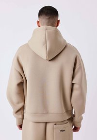 Felpa oversize beige con cappuccio ampio, polsini e fascia in vita a costine. Presenta una tasca frontale e una texture sottile. Vista posteriore.