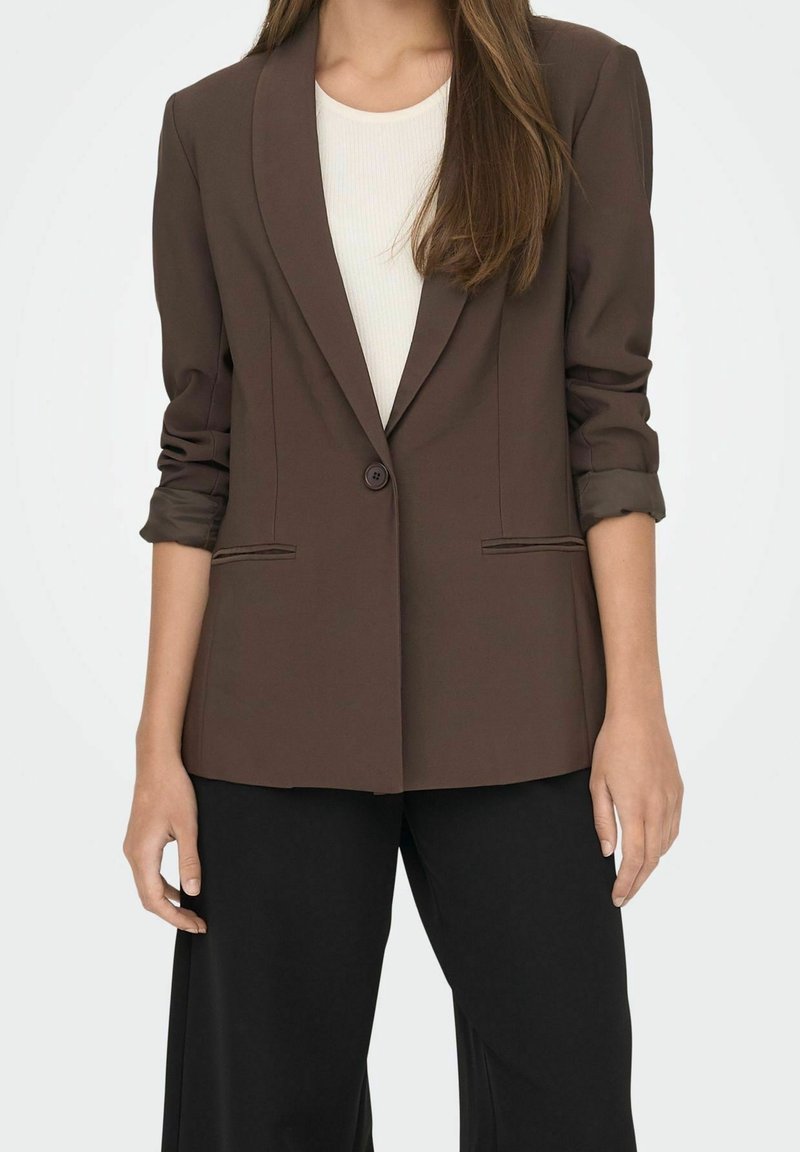 Bruine blazer van lichtgewicht materiaal, met een enkele knoopsluiting, opgerolde mouwen en zijzakken. Gecombineerd met een aansluitend wit topje en zwarte broek.