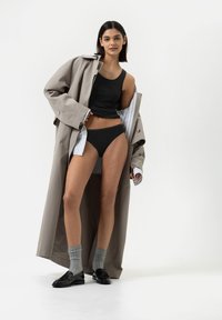 Schwarzes Tanktop, schwarzer Bikini und graue Socken kombiniert mit schwarzen Loafers. Trägt einen beigen Trenchcoat mit hell gestreiftem Futter, offen vorne.