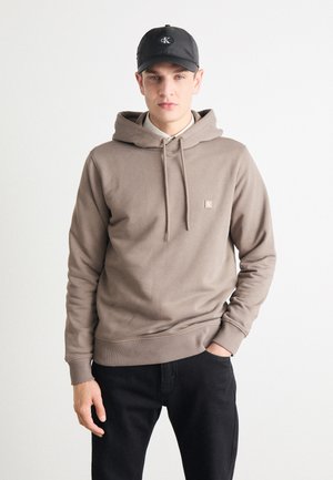 Sudadera marrón con capucha ajustable, que presenta un pequeño logo en el pecho. Combinada con jeans negros y una gorra negra. Textura de tela suave.