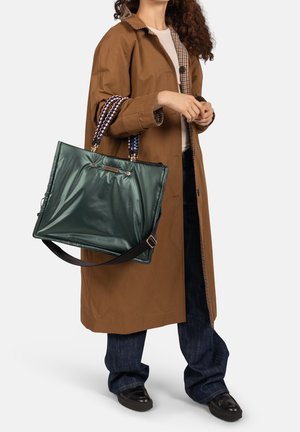 Vrouw met een bruine jas, blauwe spijkerbroek, zwarte schoenen, en ze draagt een grote donkergroene handtas met meerkleurige gevlochten handvatten.