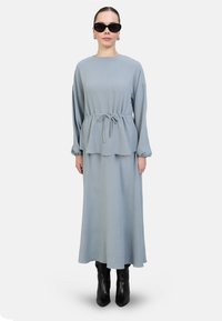 Helles blaues Kleid mit langen Ärmeln, taillierter Taille und A-Linie-Silhouette. Kombiniert mit schwarzen, knielangen Stiefeln und übergroßen schwarzen Sonnenbrillen.
