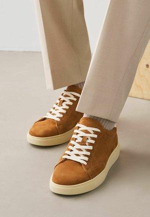 Brune ruskindssneakers med hvide snørebånd, båret med beige bukser og lyse ribstrikkede sokker på et neutralt gulv.