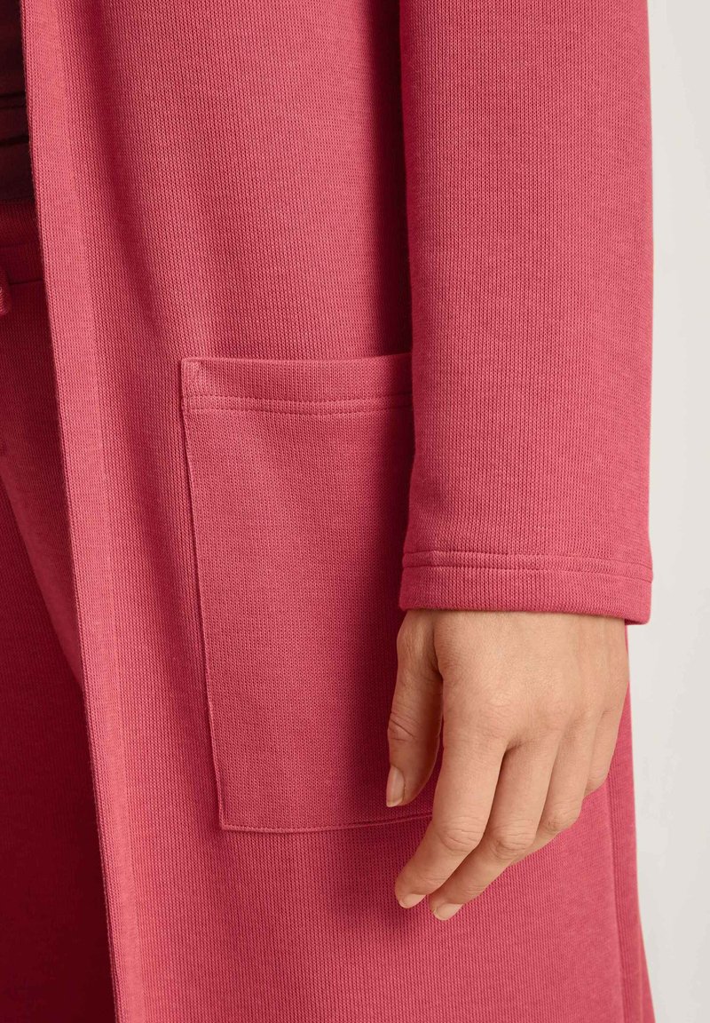 Langer rosa Cardigan mit geripptem Textur, ausgestattet mit einer Seitentasche und schmalen Ärmelabschlüssen, vor einem hellen Hintergrund gezeigt.