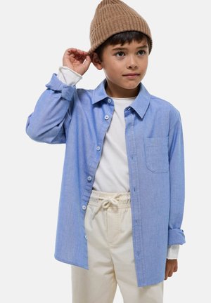 Chemise boutonnière bleu clair avec une poche, portée par-dessus une chemise blanche à manches longues. Associée à un pantalon beige à cordon de serrage et un bonnet brun en côtes.