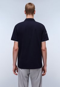 Mörkblå pikétröja med korta ärmar, vit kant vid kragen och ärmsluten. Bakre vy visar slät tygkvalitet och rena designlinjer.