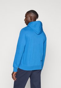 Μπλε φούτερ με κουκούλα, τσέπη καγκουρό και ρυθμιζόμενη κουκούλα. Κατασκευασμένο από απαλό ύφασμα, με υφή, και εφαρμοστό στα μανίκια και το τελείωμα. Συνδυάζεται με σκούρο παντελόνι.