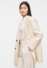 Μπεζ υπερμεγέθη σακάκι, υφασμένο με υφή, κλείσιμο με ένα κουμπί, συνδυασμένο με ροζ ανοιχτόριχτο ριμπ πουκάμισο και παντελόνια σε ίδιο χρώμα.