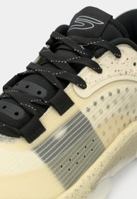 Κοντινή λήψη μπεζ και μαύρου αθλητικού παπουτσιού με κορδόνια με σχέδιο, ενδιάμεση σόλα με στίγματα και υφασμένο πλαϊνό πάνελ με μικρές οπές αερισμού.