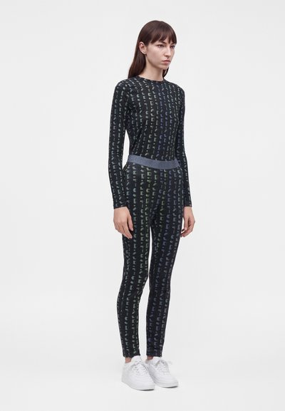 Femme debout portant un haut noir à manches longues à motifs et des leggings assortis avec des baskets blanches sur un fond blanc.