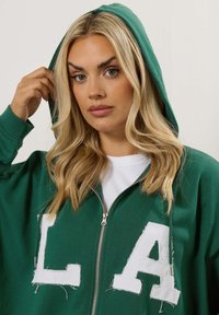 Felpa verde con zip e logo "LA" bianco sbiadito sul davanti, dotata di cappuccio e una texture di tessuto morbido. Il modello tiene il cappuccio.